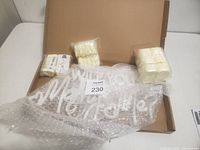 Wide view of unopened brown box with the Marry Me light-up sign partially wrapped in bubble wrap laid inside along with three packs of white decorative petals on the left and right sides.