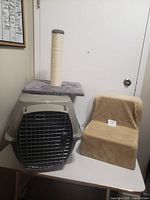Photo showing gray plastic cat cage with metal door, gray carpeted platform on top with scratching post, and beige carpeted stairs.