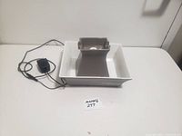 Photo shows ceramic cat water dish fountain on a white surface with attached black power adapter. The dish has a square base with a raised gray center. Good condition with no visible cracks. Power adapter appears intact.