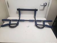 Front view of metal exercise pull up bar with blue foam padded grips laid flat against a closed white door, showing the full construction and grip placement.