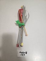 Knitting utensils including several knitting needles and a needle gauge tool, bundled together with a green twist tie, arranged on a white surface.