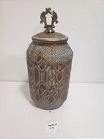 Photo of the vintage antique jar standing upright, showing detailed geometric patterns and metal lid with ornamental handle