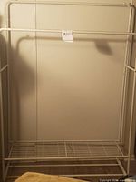 Full view of the white metal coat rack showing the hanging rod and bottom wire grid shelf against a wall.