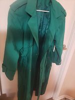 Green women's spring coat hanging on a white hanger on a door. Visible details include shoulder epaulets, belted waist, collared neckline, and button/snap front closure.