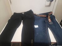 Two pairs of women's pants, black and blue denim, displayed on floor against white door. Both have belts.