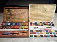 Open wooden watercolour paint box with multiple dried paint pans, paint mixing areas and two brushes, painted lid interior showing artist's tools and imagery.