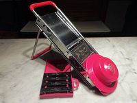 Full view of Bron Coucke mandoline with red finger guard, fold-out leg, and closed julienne blade case on countertop