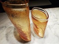 Two amber art glass vases together, showing taller vase with irregular shape and controlled bubble texture and shorter, curved vase with similar texture and amber color.