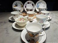 Full set of nine fine bone china tea cups and matching saucers, all with different floral patterns arranged on marble surface.