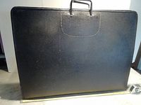 Closed black art portfolio case with a handle, textured surface visible, approximately 26x20 inches in size.