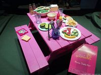 Bright pink picnic table for 18-inch dolls with picnic accessories arranged on table including plates with plastic food, glasses, and utensils.