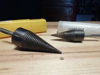 Close-up of the two spiral metal wood splitters with tapered, threaded cone tips sized approximately 42mm and 32mm, placed on a wooden surface with their cases behind them, one yellow and one white translucent.