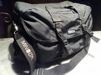 Side view of the Tenba black camera bag showing shoulder strap with Minolta logo, heavy duty fabric, and buckle straps on front flap.