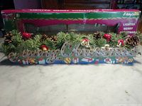 Photo showing the full holiday sign in original packaging, decorated with artificial greenery, pine cones, and red berries.