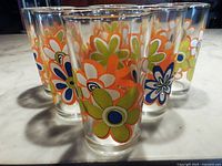 Six clear glass tumblers with vintage-style flower power floral patterns displayed on a marble countertop