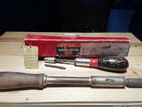 Photo showing two vintage spiral ratchet screwdrivers placed on a wooden surface: one with red/black handle and original red box, and another wooden handled screwdriver without box.