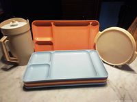 Beige Tupperware juice jug with handle and matching covered dish with lid beside stack of multi-colored picnic trays.