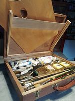 Open wooden artist box showing interior with paint tubes, brushes and small jars.
