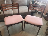 Two vintage teak chairs with pink upholstered seats placed side by side on a carpeted floor in a living room setting.