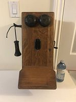 Full front view of antique wooden crank telephone mounted on wall with receiver and crank, showing size relative to water bottle.