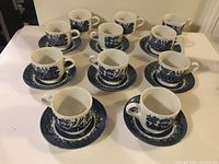 Eight Blue Willow cups with matching saucers arranged neatly on white table with three additional cups placed among them.