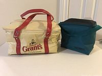 Front view of the cream Grants Scotch Whiskey cooler bag with red handles next to the teal Thermos lunch bag
