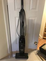 Full view of black Eureka The Boss stick vacuum with power cord wrapped on the back, standing upright in front of a door.