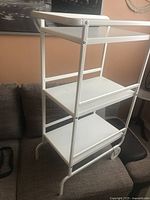 Side angle view of the white metal trolley showing three shelves and two legs without wheels.