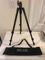 Full view of black Slik tripod extended on table with carrying case in front.