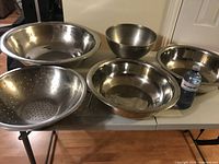 Top-down angled view of the five stainless steel bowls laid out on table including a large mixing bowl, smaller bowls, and a colander, shows size and condition of each.
