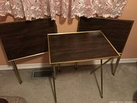Three vintage TV trays with dark wood grain tops and gold trim, standing folded against a peach-colored wall beneath lace curtains.