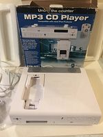 White MP3 CD player with iPod dock and control buttons, shown with original blue box in background