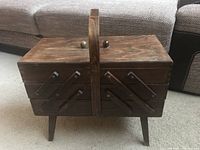 Closed view of the wooden accordion-style sewing box with legs, showing dark wood finish and central handle.