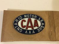 Oval metal plate badge showing the CAA logo in red on white, surrounded by blue with raised silver text "ONTARIO MOTOR LEAGUE AAA." Mounted on brown paper backing.
