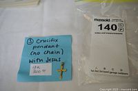 Crucifix pendant displayed on a blue note with handwritten details and a price tag, placed next to a MaxSold bag with lot number.