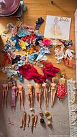 Overview photo showing assorted Barbie dolls, detached limbs, and numerous doll clothing pieces spread on floor and carpet.