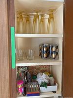 Full view of cabinet shelf containing amber pilsner beer glasses, clear short tumblers, black and gold tumblers, assorted stir sticks, and other barware items.
