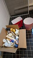 View of a cardboard box filled with assorted personal care products including bottles of shampoo, conditioner, body lotion, toothpaste, and soaps. Visible items vary in size and color, mostly with white or yellow caps.