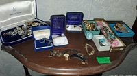 Wide view of assorted costume jewelry spread on a wooden table including watches, earrings, rings, pendants, and bracelets in boxes and loose.