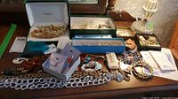 Overview of entire costume jewelry collection on wooden table showing various items including boxed jewelry, loose chains, beaded and pearl necklaces, and earrings.
