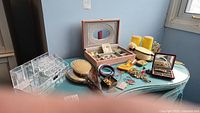 Various costume jewelry items, pink jewelry box, acrylic holder, two vanity brush and mirror sets, and other small accessories laid out on a table.