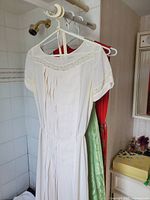 Three dresses hanging on white and pink hangers, showing front and side views. Dresses are white with lace trim and pleated details.