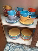 Top shelf showing eight ring-handled cups in yellow, grey, turquoise, purple, red on matching saucers