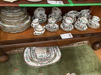 Front view showing four teacup and saucer sets, creamer, sugar bowl and additional stacked soup bowls and plates on wooden shelf.
