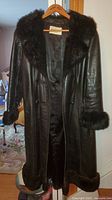 Front full-length view of the black leather coat showing faux fur trim on collar, cuffs, and hem.