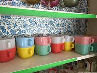 Photo of mugs stacked in pairs, showing colors white/red, blue/yellow, pink/green, white/red, yellow, pink/green stacked on a shelf with floral wallpaper background.