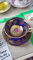 Top angle showing cobalt blue teacup with intricate gold designs and pink cabbage rose inside, paired with matching saucer below.