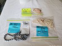 Three sealed Ziploc bags on white fabric: one containing a grey pearl necklace, one with a white double-strand necklace, one with freshwater pearl set and sticky notes