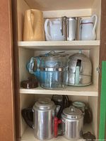 Cupboard shelves with percolators, pitchers, and teapots showing variety of materials and styles