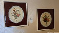 Two framed floral prints hung on a patterned wallpaper wall, each with an oval floral bouquet print, side by side.